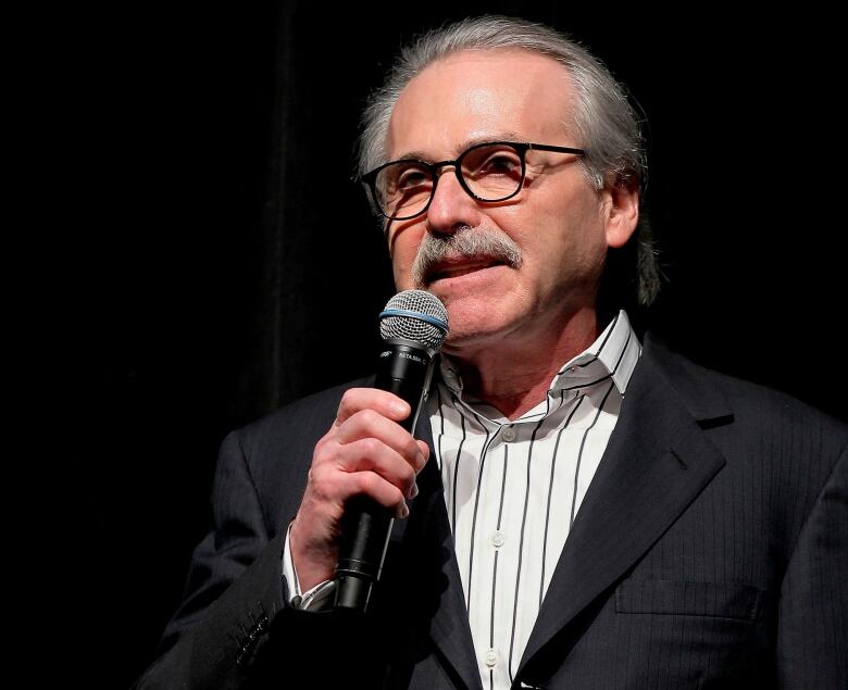 A mustached man in a suit holds a microphone in front of a darkened background.