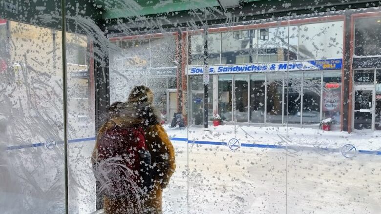 A woman in a coat stands with her back to the camera. She is inside a bus shelter and seen through a window covered in frost.