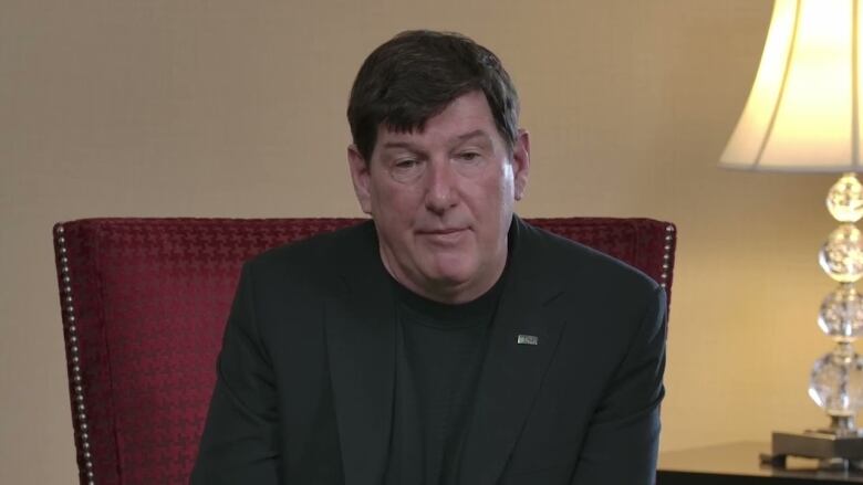 A middle-aged white man wearing a dark suit jacket over a black sweater. He has black hair.