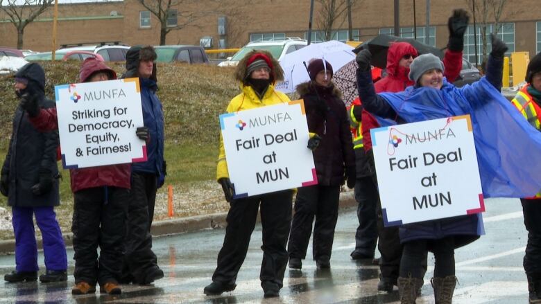 People wearing winter clothing stand in the rain while holding picket signs that say things like 