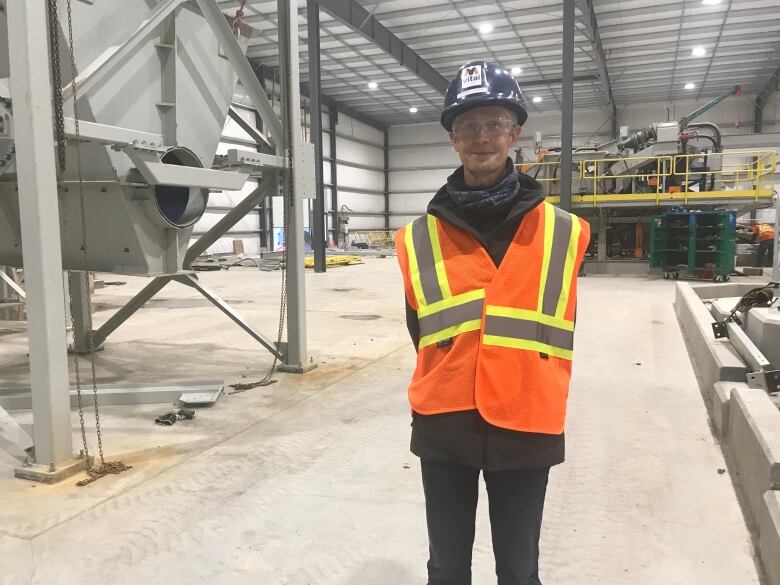 A man wearing a hardhat, safety glasses and an orange and yellow safety vest stands in an industrial processing plant. 