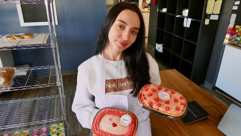 A girl with long dark hair stands in front of a counter wearing a white apron. In each hand she has a clear containers that contain baked goods.