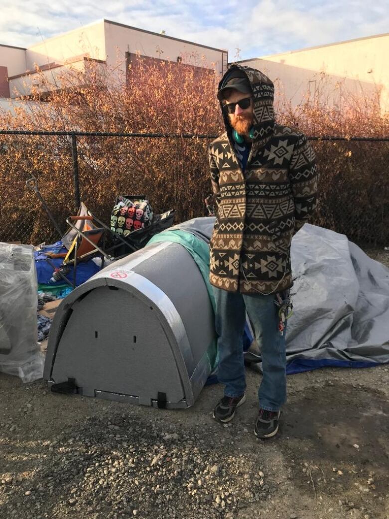 A man in a warm coat with hood stands outside of a grey, tube-shaped foam structure placed up against a fence in a designated lot in Kelowna.