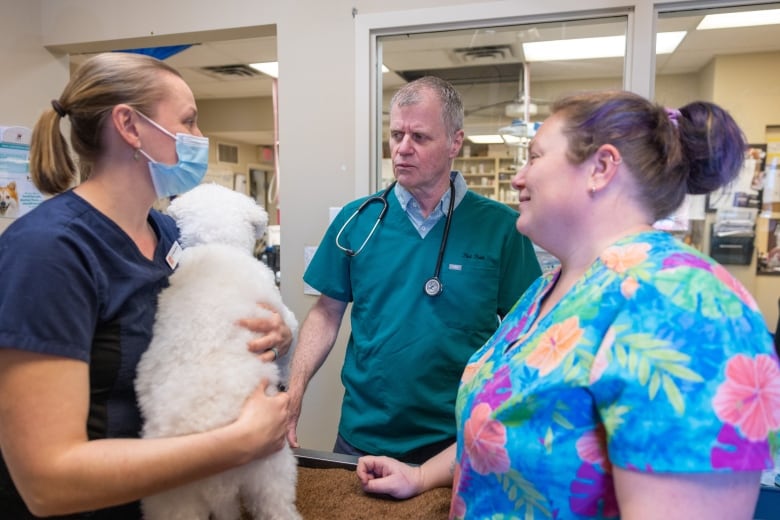 A man stands in between two women. He is wearing green scrubs.