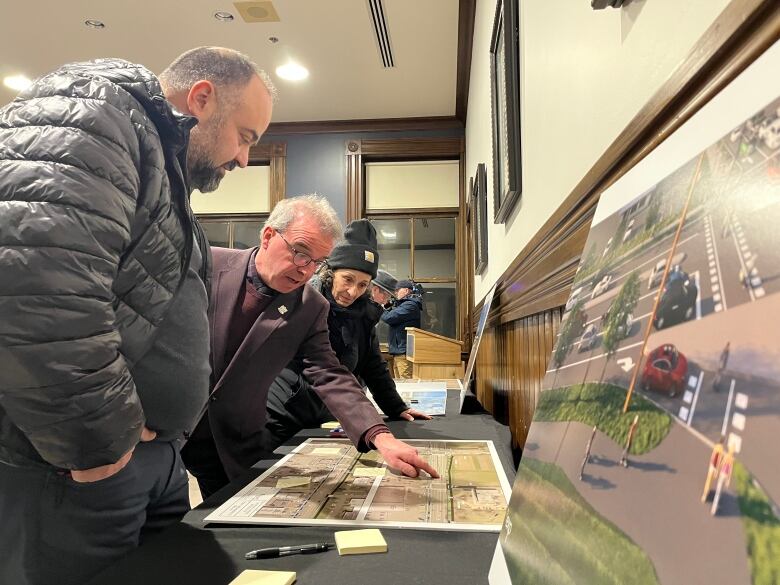 Three people look at a billboard on a long table, showing maps of a street. 