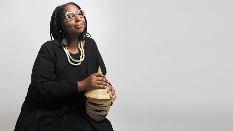 Black woman in a black dress, large beaded necklace and funky glasses holds an African basket in her lap.