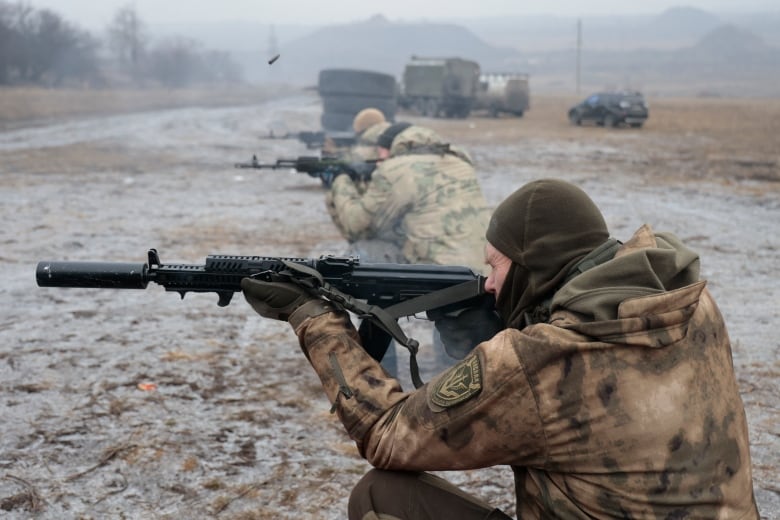 Soldiers in camoflauge kneel in a line outdoors while using automatic weapons. Military vehicles are visible in the background and the ground is covered in a dusting of snow or frost.