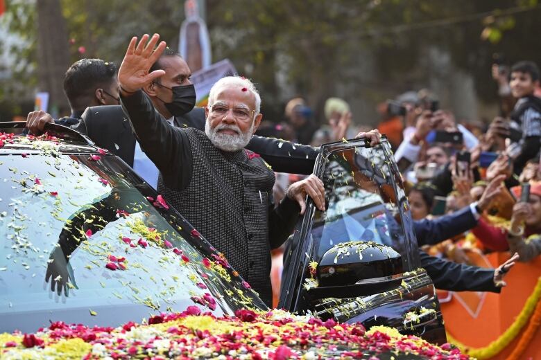 Indian Prime Minister Narendra Modi exits a vehicle and waves to supporters. There are two security officers behind him and there is a crowd in the background. The car is covered in flower petals. 