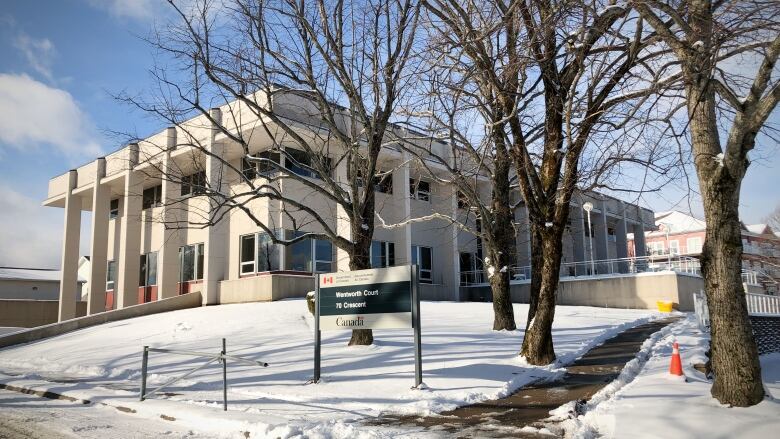 A two-storey building with large columns on the outside is seen among bare trees in winter with snow on the ground.