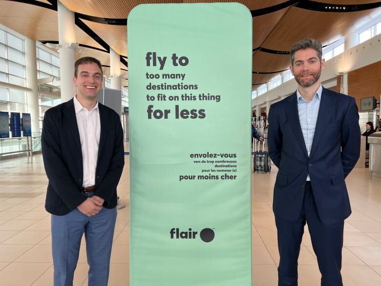 Two men in suit jackets pose next to a Flair Airlines sign in an airport.