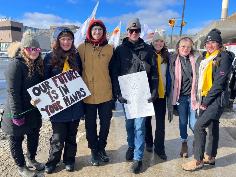 Six people wearing winter clothing stand on a sidewalk. One is holding a sign which reads our future is in your hands. 