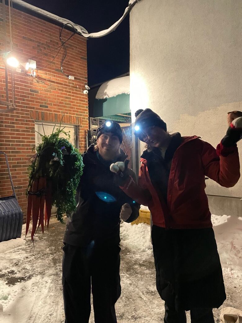 Jasmin Cartier and Grace Poland pose with carrots and garlic in front of a dumpster, wearing headlights on a recent dumpster diving excursion. 