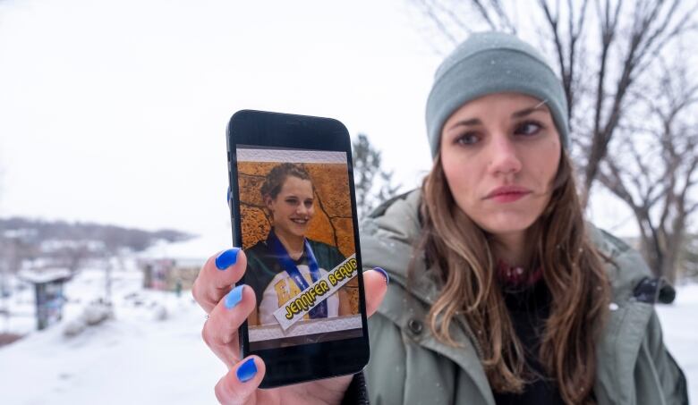 A woman wearing a tuque and parka stands outside. She's gazing at a photo of her teenaged self, shown smiling and wearing a sports uniform. 