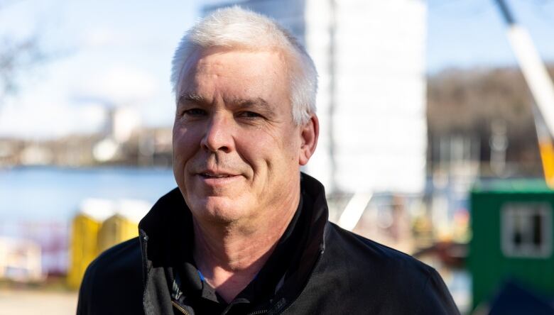 A white man with white hair stands in front of a lake, looking directly into the camera.