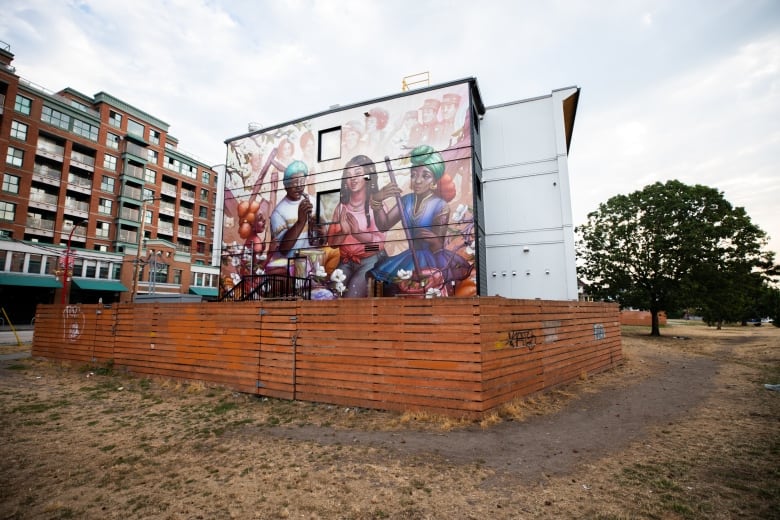 A mural in Vancouver's Hogan's Alley surrounded by fencing features pictures of Black people performing various activities. Highrises can be seen to the left.