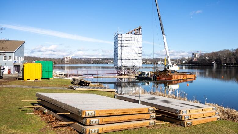 The tower in cladding and scaffolding is seen from the shoreline, with planks of wood visible on the grass in the foreground.