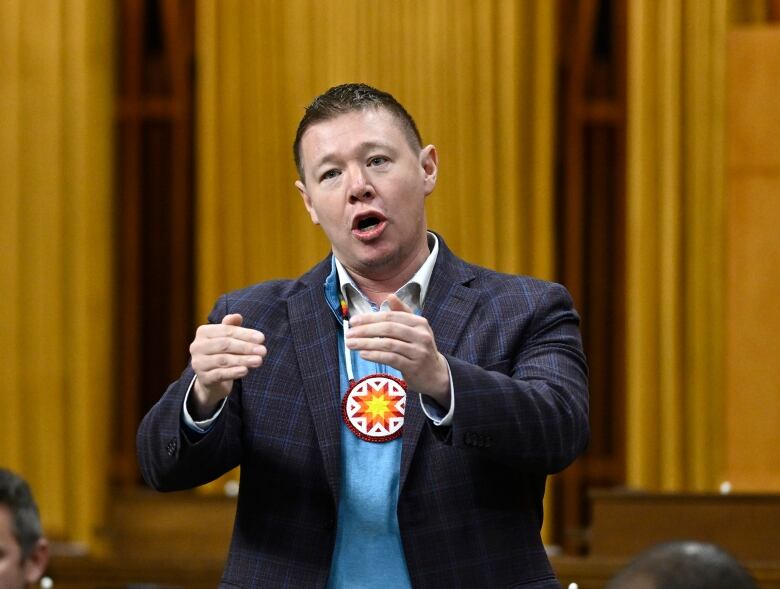 A man in a blue suit with an Indigenous symbol on a necklace gestures with his hands as he speaks.