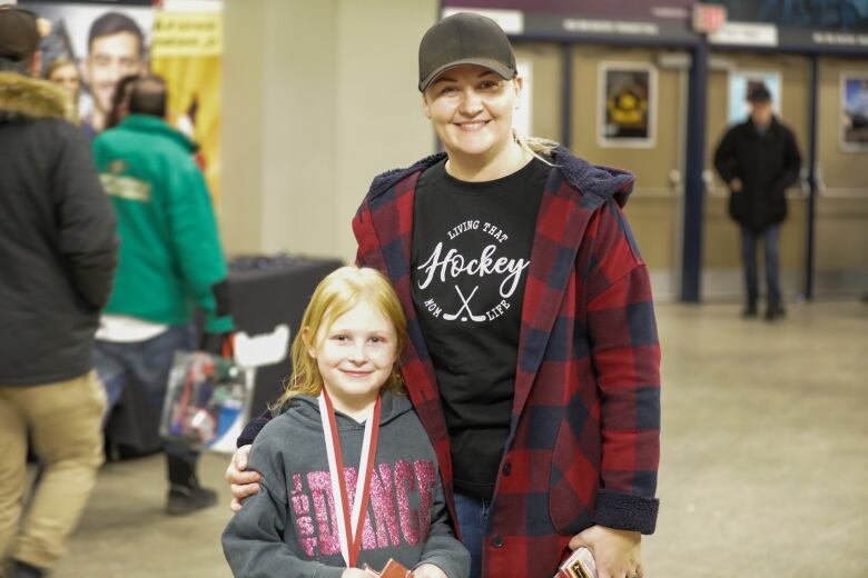 A woman wearing a black baseball cap, flannel jacket and black shirt that reads, 'Living that hockey mom life,' stands with her arm around a little girl with strawberry-blonde hair. The girl is wearing a charcoal grey hoodie that reads, 'Just dance,' and has a lanyard hanging from her neck. Both of them are smiling toward the camera. They are standing in the concourse of a hockey arena, with people walking past them in the background.