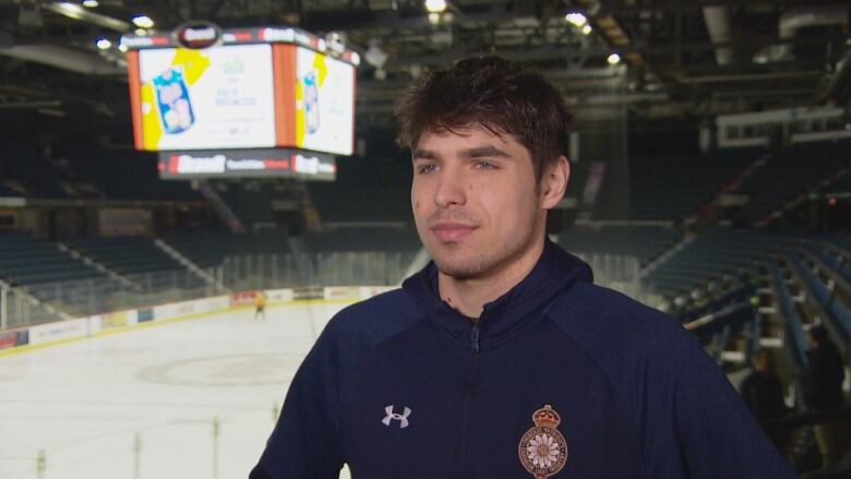 A young man with black hair and stubble is wearing a navy blue zip-up hoodie, with the Underarmour and Regina Pats logo on the chest. He is standing in a near-empty hockey arena. Behind him is a jumbotron and the ice. 