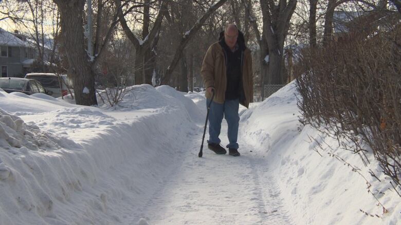 A man using a cane is walking down a sidewalk that is covered in snow.