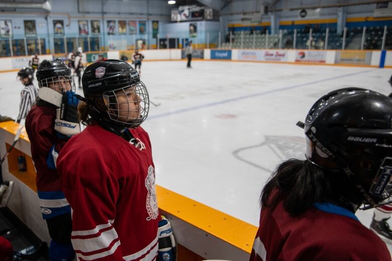 Hockey players stand on the bench, they're turned toward the action on the ice. 