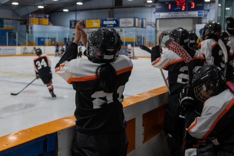 Young hockey players stand up on the bench, clapping their hands together and cheering on their friends on the ice. 