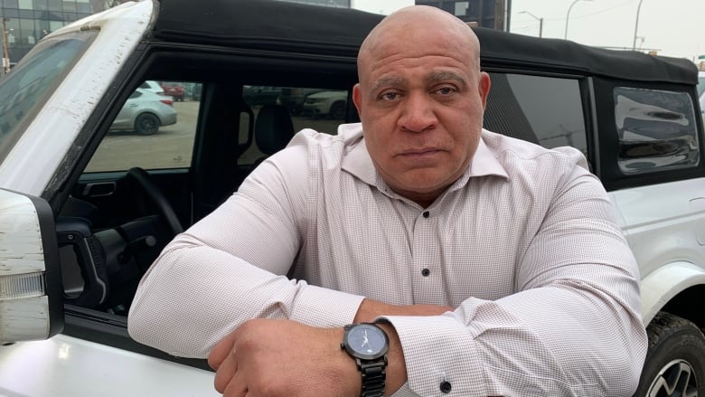 Wil Brookins, a Black man in a white dress shirt, stands beside the open door of his jeep outside.  