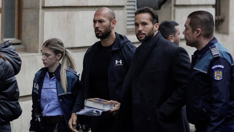 Two men in dark clothing are escorted by police officers into a court building.