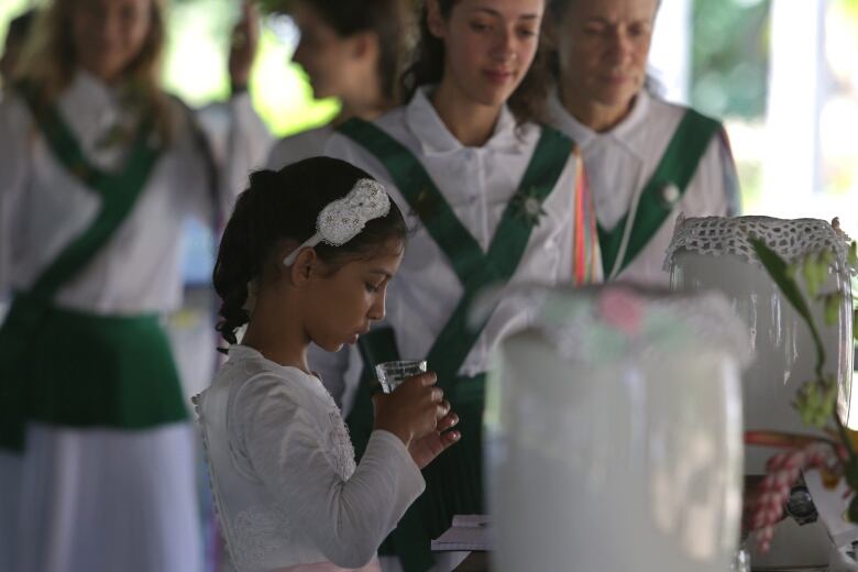 A 9-year-old girl in white kneels at an altar in Brazil sipping a tea.