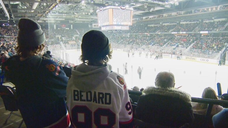 Two boys wearing hockey jerseys stand in a packed hockey arena. They overlooking the lower bowl of the arena and the ice. In the background the stadium seats are full with people.