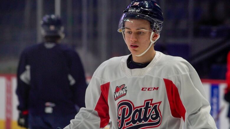 A hockey player is looking toward the ice. He is wearing a white practice jersey that reads, 'Pats,' on the front. He is wearing a navy blue helmet, that has a white chinstrap and a visor, and features the number 98 on the front.