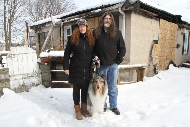 A woman, man and dog stand outside a home with fire damage and boarded up windows and doors.