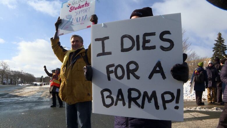 Strikers standing on a roadway holding signs with their arms raised. The sign in front reads I dies for a barmp. 