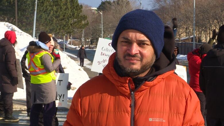 A man wearing a blue winter hat and an orange jacket standing in front of striking workers. 