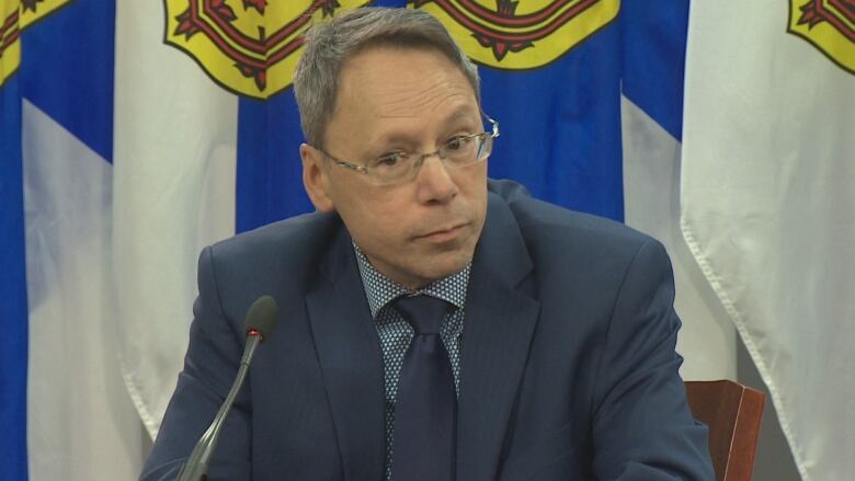 A man with glasses and gray hair sits at a podium in front of Nova Scotia flags.