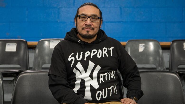 A man sits in a hockey arena for a portrait, he's wearing a shirt that says Support Native Youth. 