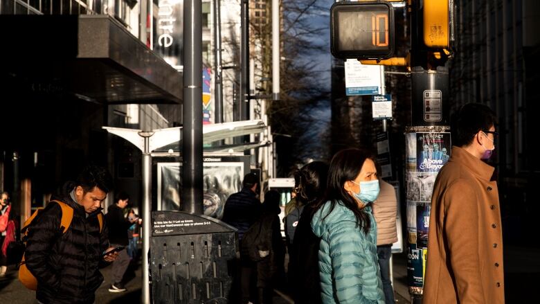 A small crowd of people walk through downtown Vancouver, a woman and a man in the foreground are wearing surgical masks. A street sign reads 