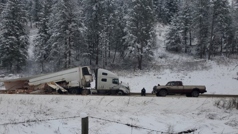 The trailer of a transport truck is smashed and its contents are lying on the highway. A pickup truck faces it, a man sits on the side of the highway.