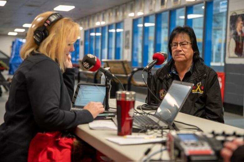 Two people sit across from each other at a table with radio-equipment set up there for a radio news special.