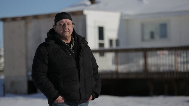 A man in a black winter jacket and jeans standing in front of a condemned home on Peguis First Nation. 