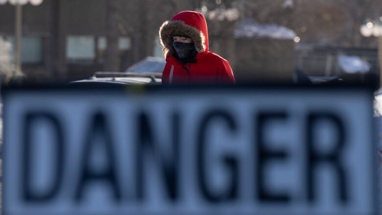 A person in a parka and scarf walks on a street while framed by a sign saying 