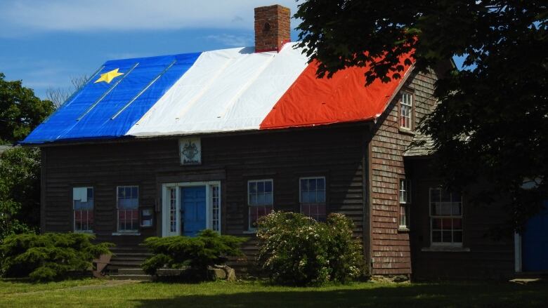 A large wooden house with a blue door and large Acadian flag draped on its roof.