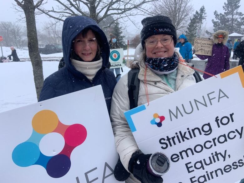 Two people wearing coats and hats hold signs with the MUNFA logo. It's snowing.