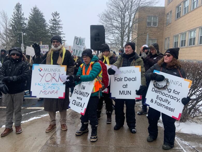 A crowd of people stand on a sidewalk, some holding signs featuring the MUNFA logo. One sign says 