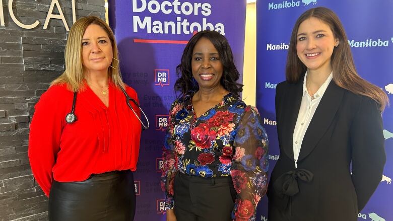 Three women pose in front of a sign that reads 'Doctors Manitoba' at a funding announcement in Winnipeg.