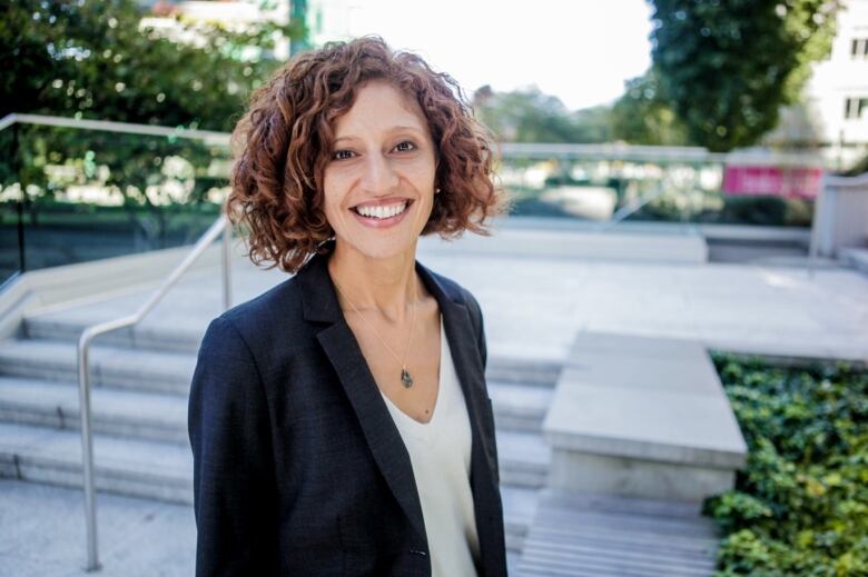 A smiling woman with reddish curly hair stands in front of some steps outside a building.  