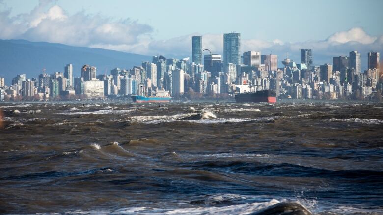 A stormy oceanfront whipped by waves with Vancouver's downtown skyline framed by mountains in the background.