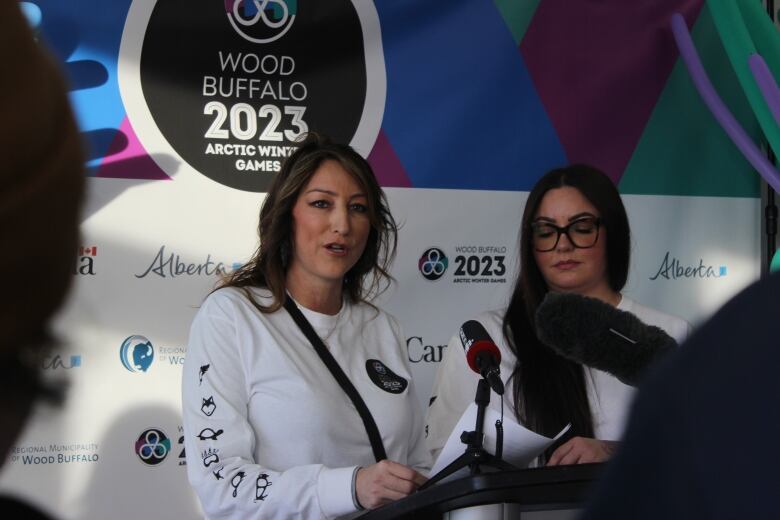 Two women in white shirts stand on a podium with a colourful backdrop. 