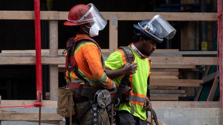 Two construction workers wearing high-vis vests are pictured in profile at a construction site. One of them is wearing a protective facemask.