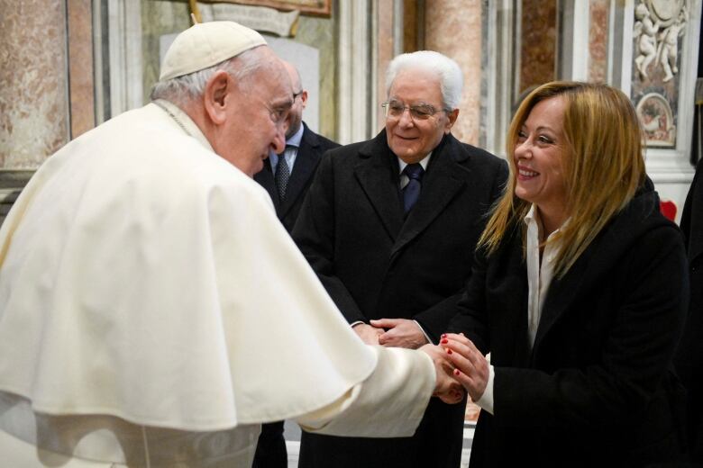 Pope wearing white regalia shakes hand of woman with long blonde hair and black jacket as a smiling man with grey hair, glasses, coat and tie watches.